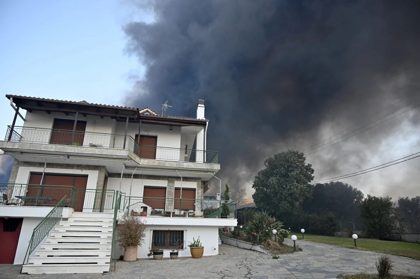 Dwupiętrowy dom z zamkniętymi okiennicami otoczony przez duże ilości gęstego, czarnego dymu unoszącego się w tle, sugerującego pożar w pobliżu budynku.