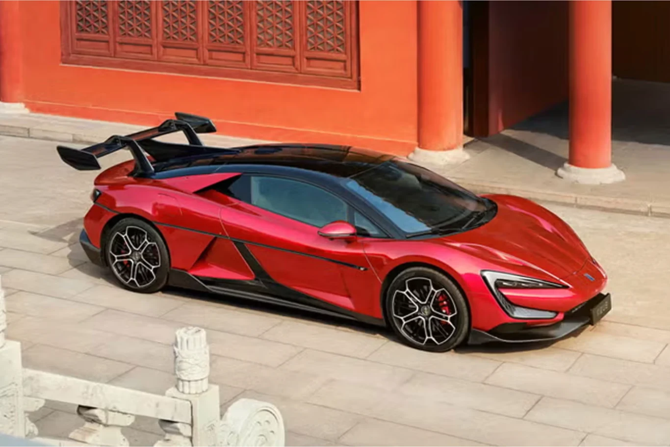 Yangwang U9 Track Edition dysponuje mocą 3019 KM. Czerwony sportowy samochód z agresywnym spojlerem oraz czarnymi elementami nadwozia, zaparkowany na brukowanym dziedzińcu przy tradycyjnym budynku z czerwonymi kolumnami i dekoracyjnym oknem.