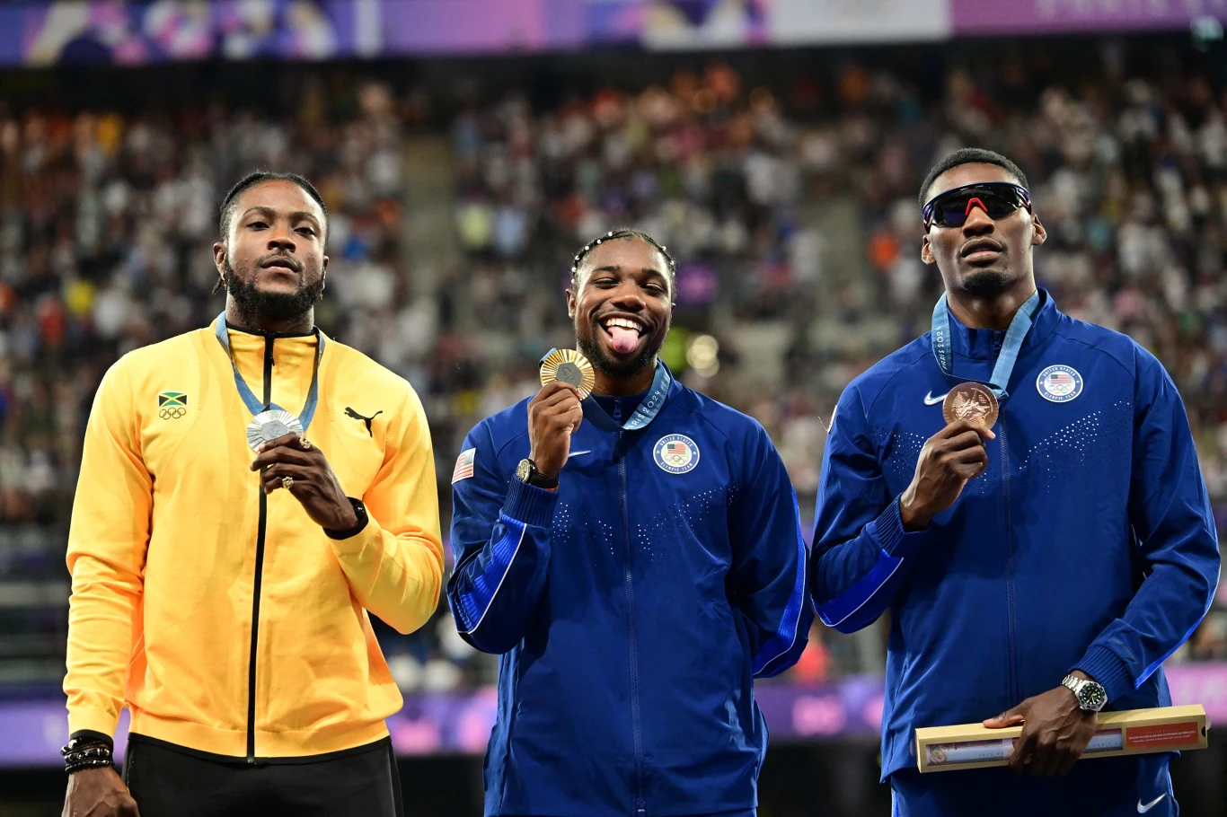 Trzech lekkoatletów stoi na podium podczas dekoracji medalowej, środkowy zawodnik trzyma złoty medal i się uśmiecha, sportowcy ubrani są w dresy reprezentacyjne Jamaiki i Stanów Zjednoczonych, w tle rozmyta widownia stadionu.