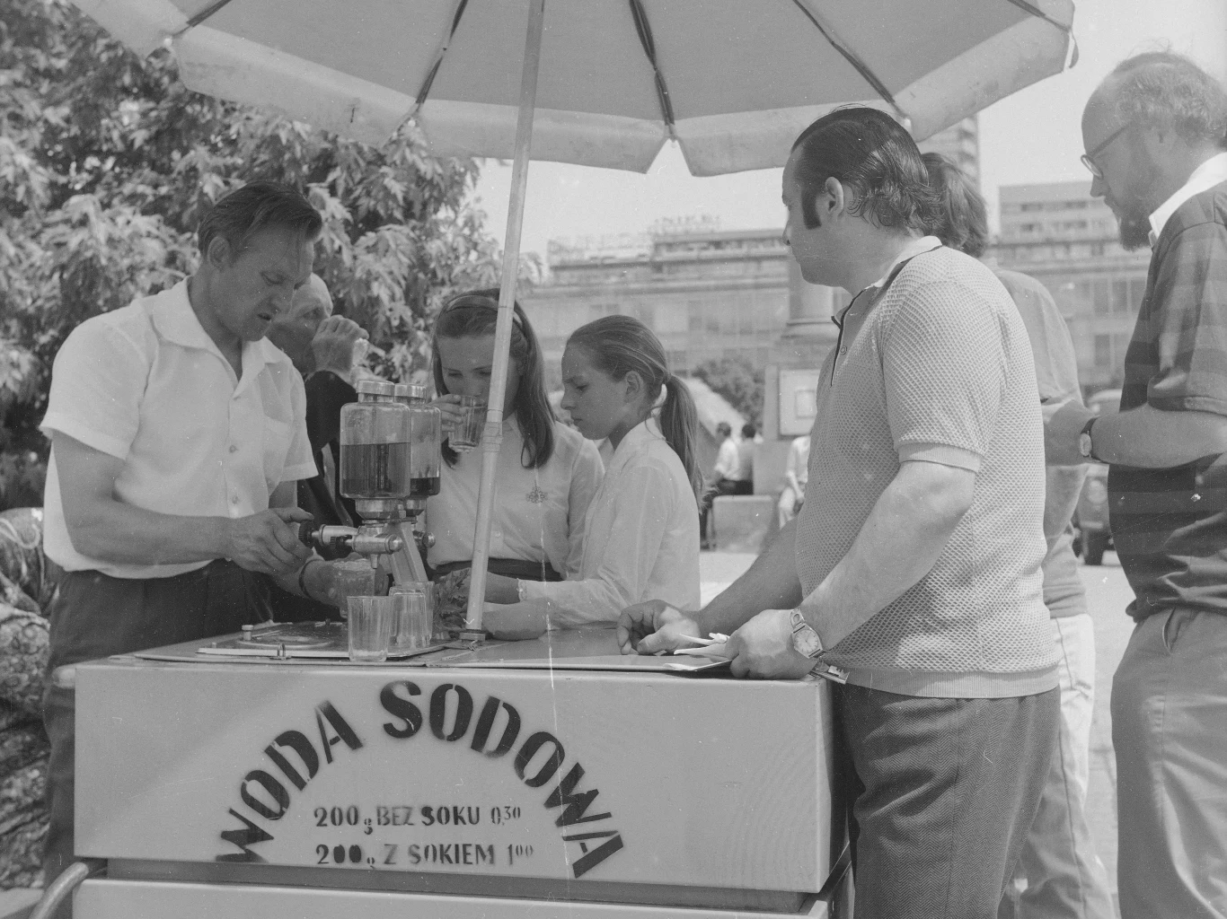 Saturator pojawił się na polskich ulicach w latach 50. XX wieku. To jeden z symboli PRL-u Stoisko z wodą sodową ulokowane na zewnątrz, sprzedawca przygotowuje napój dla dwóch dziewczynek, obok stoi dwóch dorosłych mężczyzn, nad całością rozłożony parasol chroniący przed słońcem, w tle widoczne drzewa i fragmenty zabudowy miejskiej.