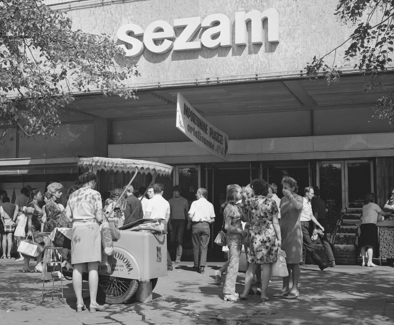 Napój serwowano w szklankach z grubego szkła, przypominających popularne wówczas musztardówki. Po wypiciu szklanka była obmywana niewielkim strumieniem wody. Grupa osób stoi przed wejściem do sklepu Sezam, część z nich rozmawia lub stoi w kolejce, z boku widoczny wózek z lodami i kobieta sprzedająca produkty.