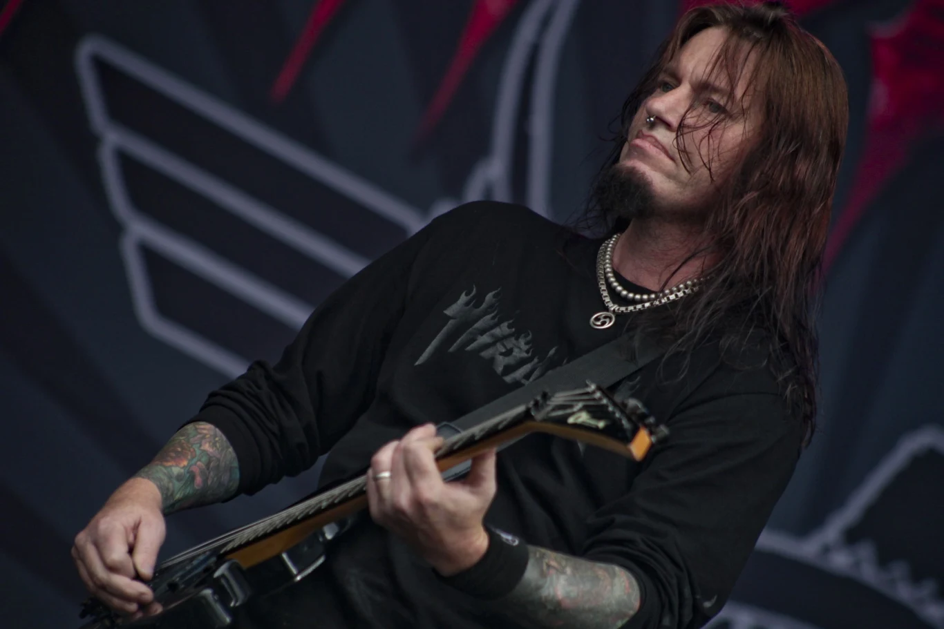 Nige Rockett w 2010 roku Mężczyzna z długimi włosami grający na gitarze elektrycznej na tle sceny koncertowej, ubrany w czarną bluzę z metalowymi aplikacjami i perłowy naszyjnik, widoczne tatuaże na obu rękach.