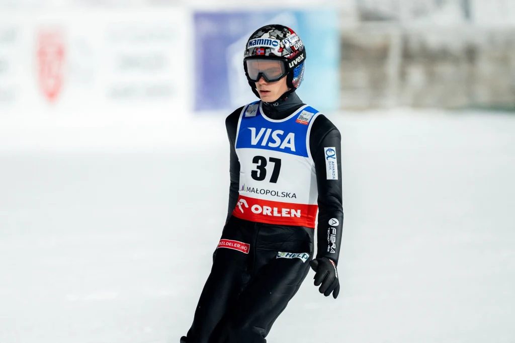 Marius Lindvik Skoczek narciarski w profesjonalnym kombinezonie i kasku, z numerem startowym 37, podczas zawodów na śnieżnym tle, z widocznymi reklamami sponsorów na stroju.