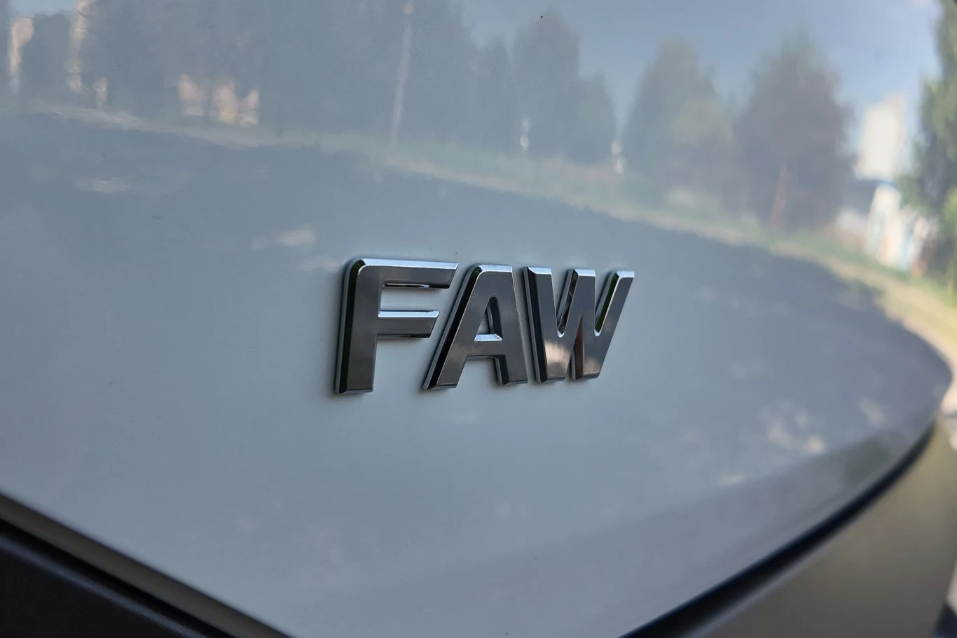 Logo FAW umieszczone na błyszczącej powierzchni samochodu, z rozmytym tłem otoczenia i delikatnymi odbiciami na karoserii.