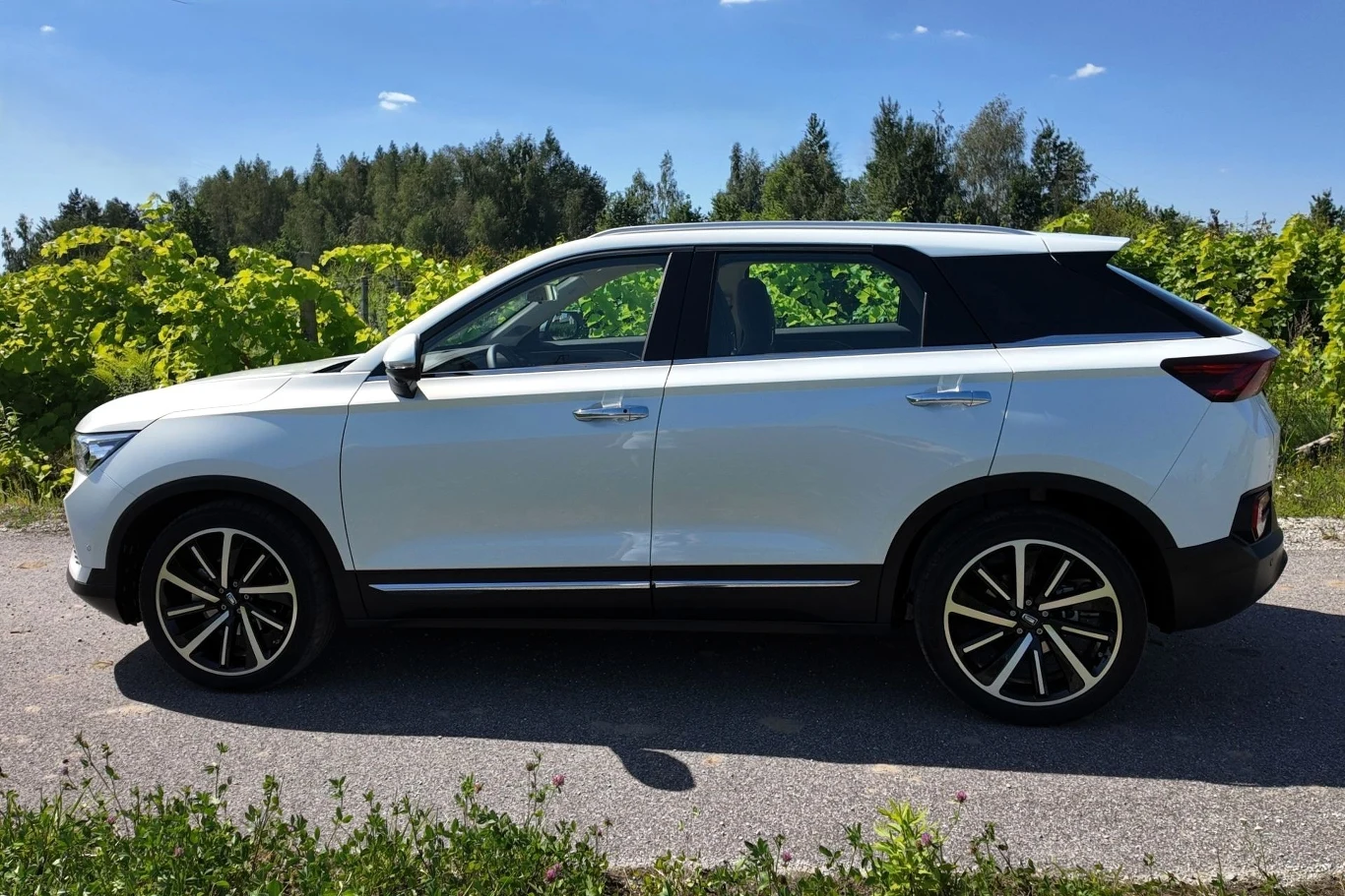 Biały nowoczesny samochód osobowy typu SUV stoi zaparkowany na asfalcie, w tle zielone krzewy oraz drzewa i błękitne niebo z pojedynczymi chmurami.