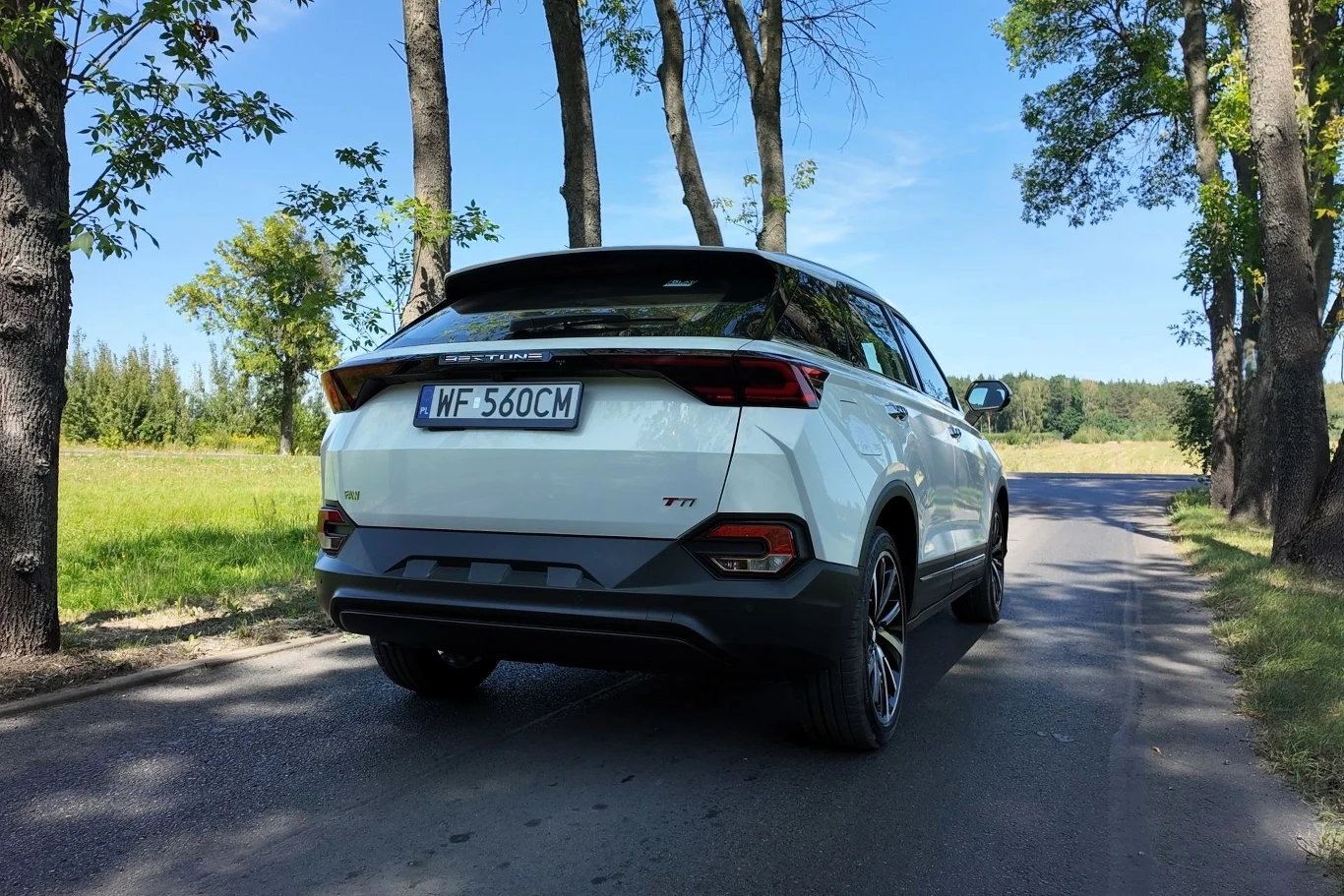 Biały SUV marki TANK 300 stoi zaparkowany na bocznej, asfaltowej drodze otoczonej drzewami i zielenią, w tle widać pole oraz rzadko rozmieszczone drzewa pod błękitnym niebem.