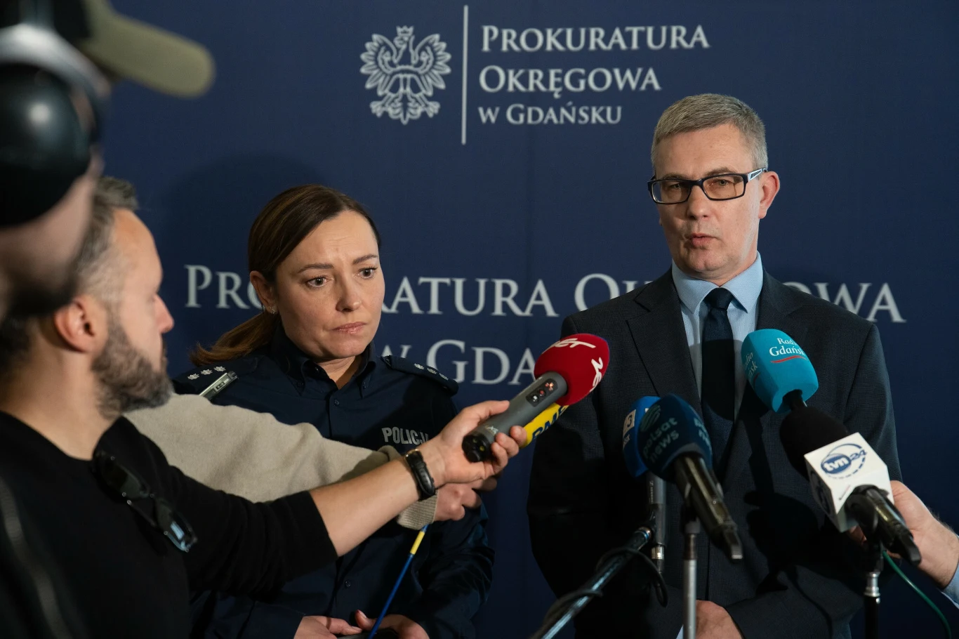 Prokuratura przedstawiła akty oskarżenia w związku z działaniem na rzecz obcego wywiadu Konferencja prasowa z udziałem funkcjonariuszki policji oraz przedstawiciela prokuratury, przed mikrofonami różnych mediów, na tle niebieskiej planszy z napisem Prokuratura Okręgowa w Gdańsku.
