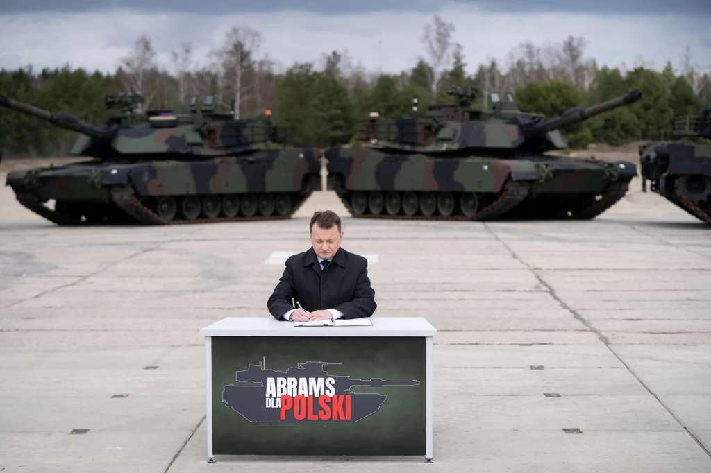 Ówczesny minister obrony narodowej Mariusz Błaszczak, podpisujący dokment umowy na pozyskanie 250 czołgów M1A2 SEPv3 dla Wojska Polskiego Mężczyzna w garniturze podpisuje dokument przy biurku z napisem „Abrams dla Polski”, w tle dwa czołgi Abrams ustawione na betonowym placu, otoczone przez drzewa.