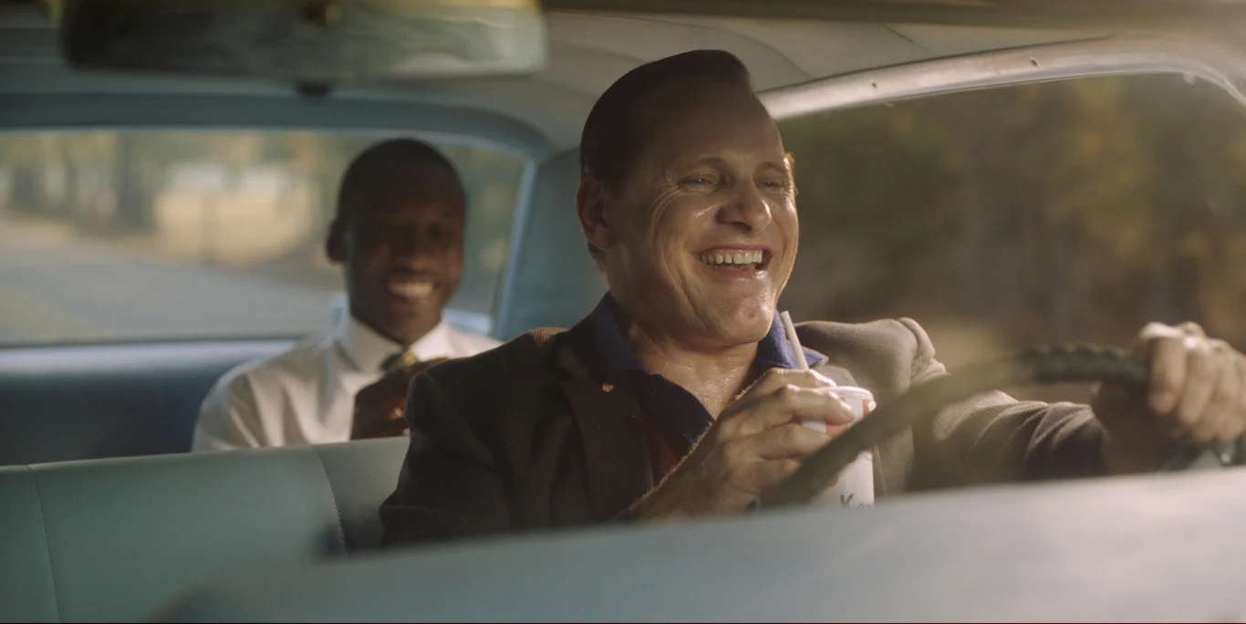 Bohaterowie filmu "Green Book" raczą się fast foodami w samochodzie Dwóch mężczyzn siedzi w samochodzie, kierowca z uśmiechem prowadzi pojazd, pasażer w jasnej koszuli siedzi z tyłu i również wydaje się być w dobrym humorze. Atmosfera jest swobodna i radosna.