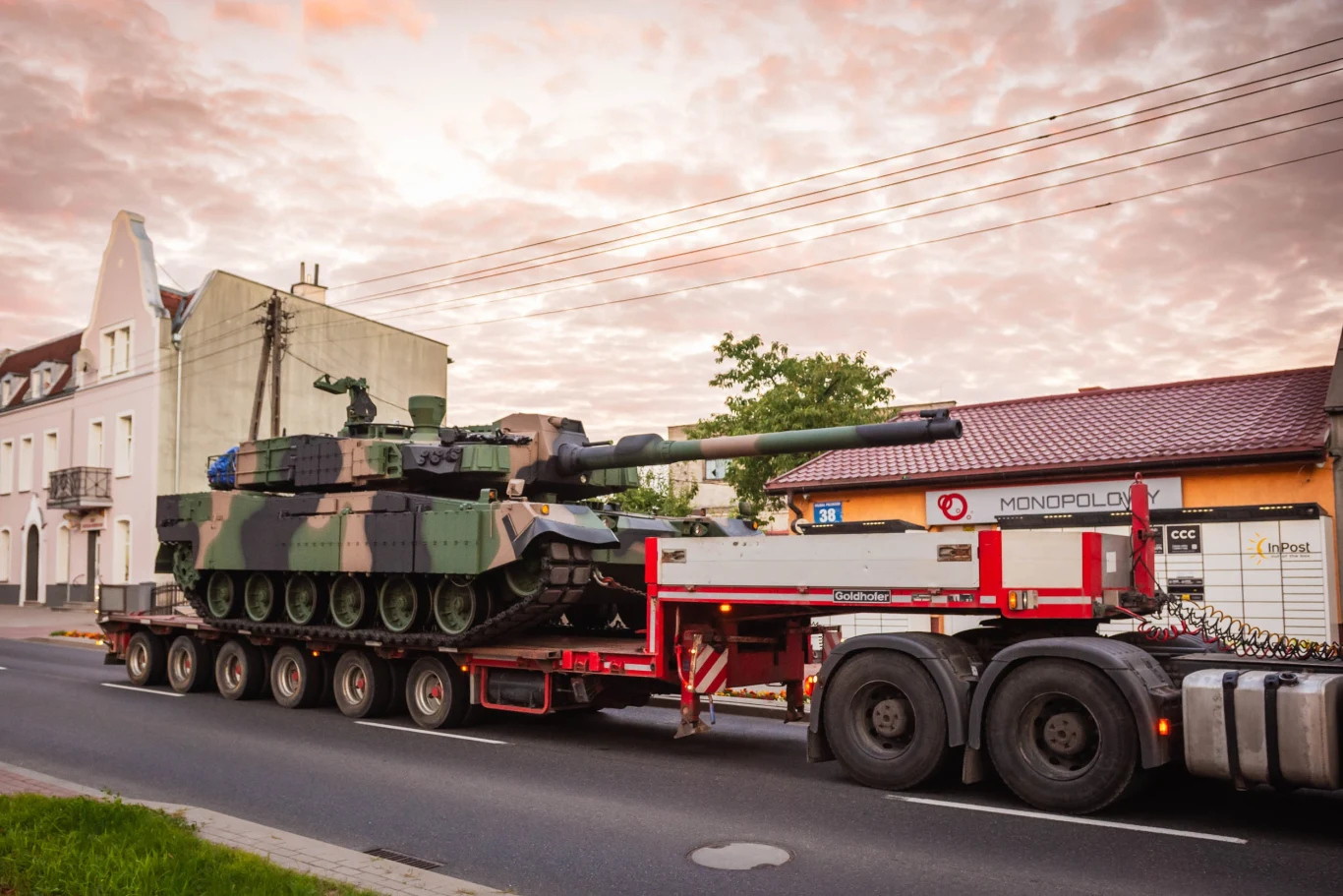 Czołg wojskowy przewożony na naczepie ciężarówki przez miasto, w tle budynki mieszkalne i punkt usługowy, nad ulicą biegną przewody elektryczne, wczesny wieczór lub poranek, niebo pokryte chmurami.