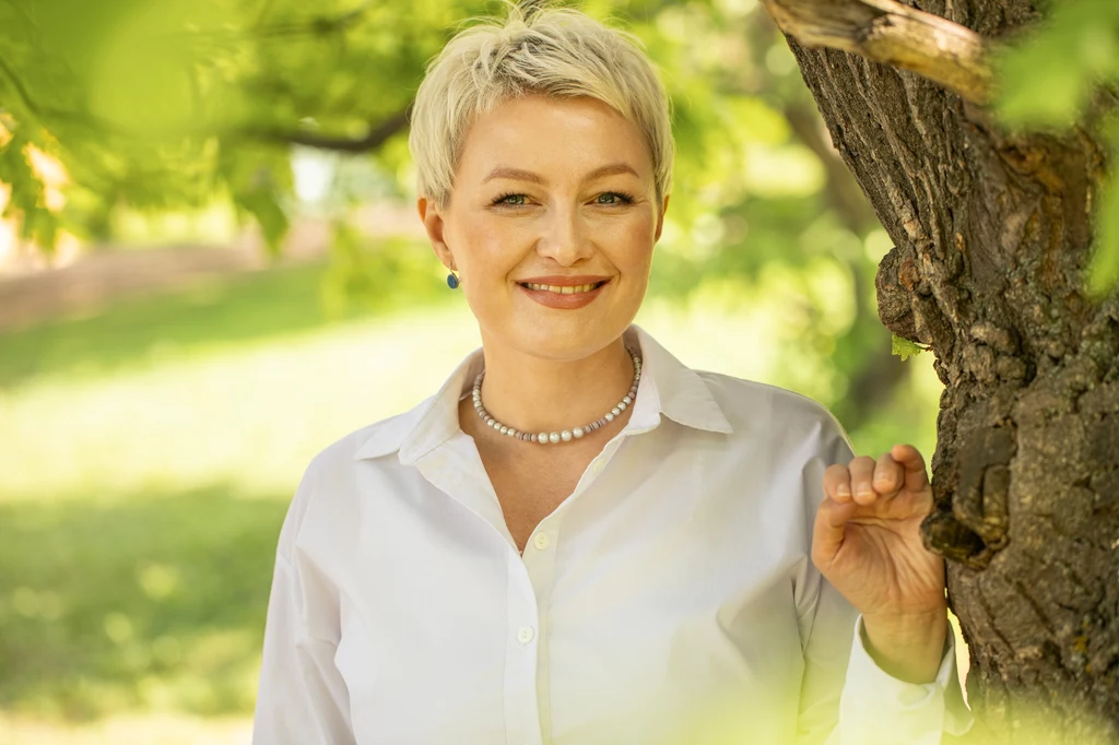 Kobieta o krótkich blond włosach i jasnej karnacji, ubrana w białą koszulę i naszyjnik z pereł, stoi przy drzewie w słonecznym otoczeniu parku, z delikatnym uśmiechem na twarzy.