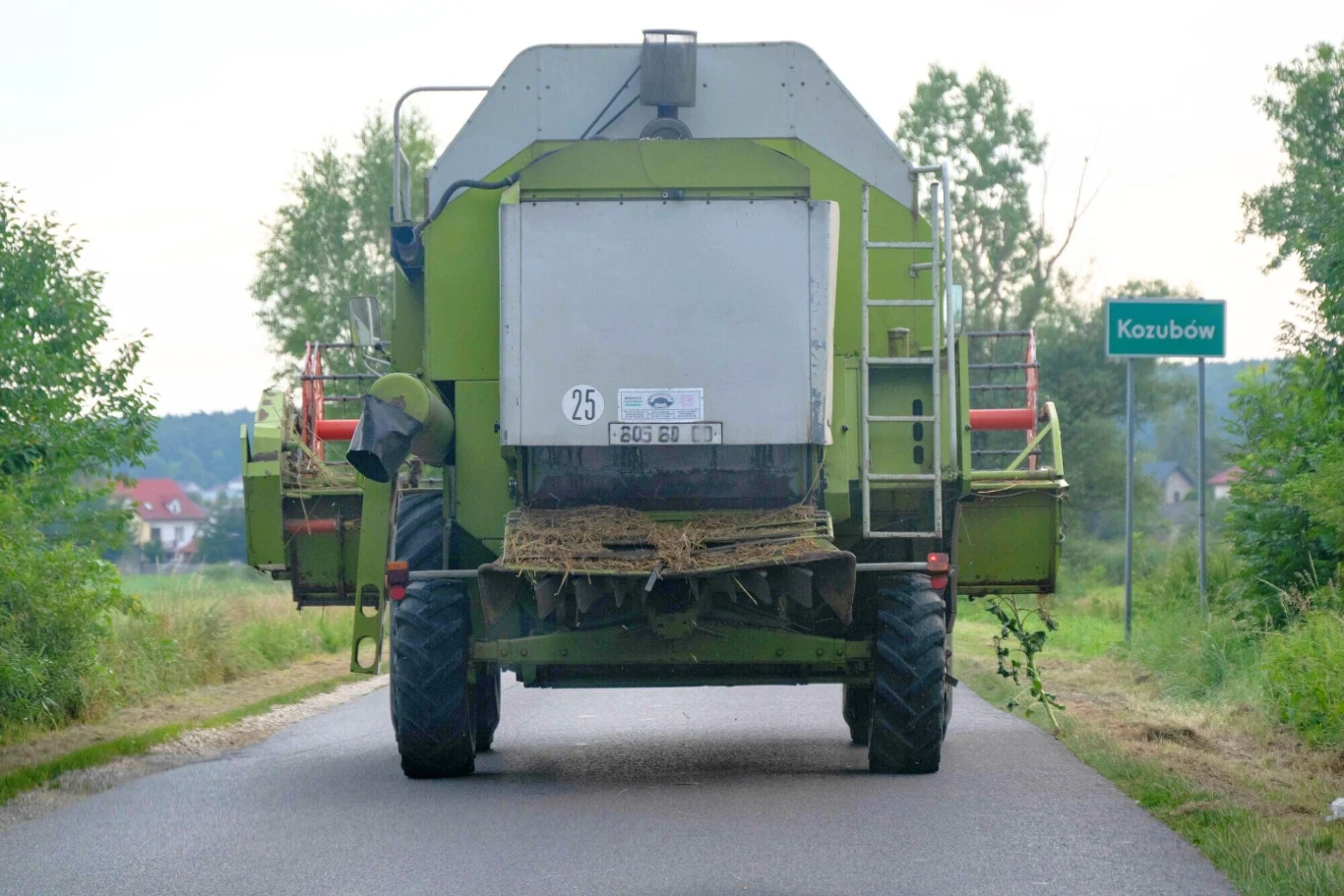 Czy kombajn na drodze powinien jechać w asyście pilota? Kombajn rolniczy jadący wąską asfaltową drogą, otoczony zielenią oraz roślinnością, wieś Kozubów wskazany tablicą przydrożną, w tle niewielkie zabudowania i drzewa.