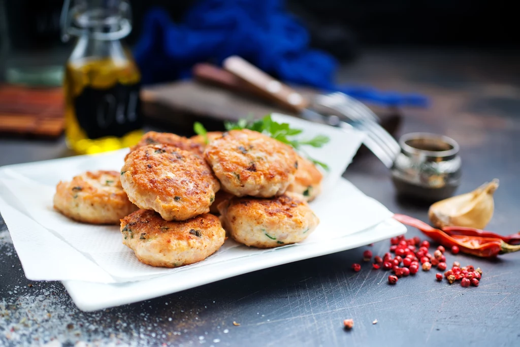 Kotleciki z mięsa mielonego podane na białym talerzu z natką pietruszki, w tle rozsypany pieprz, papryczki chilli, czosnek oraz butelka oliwy.