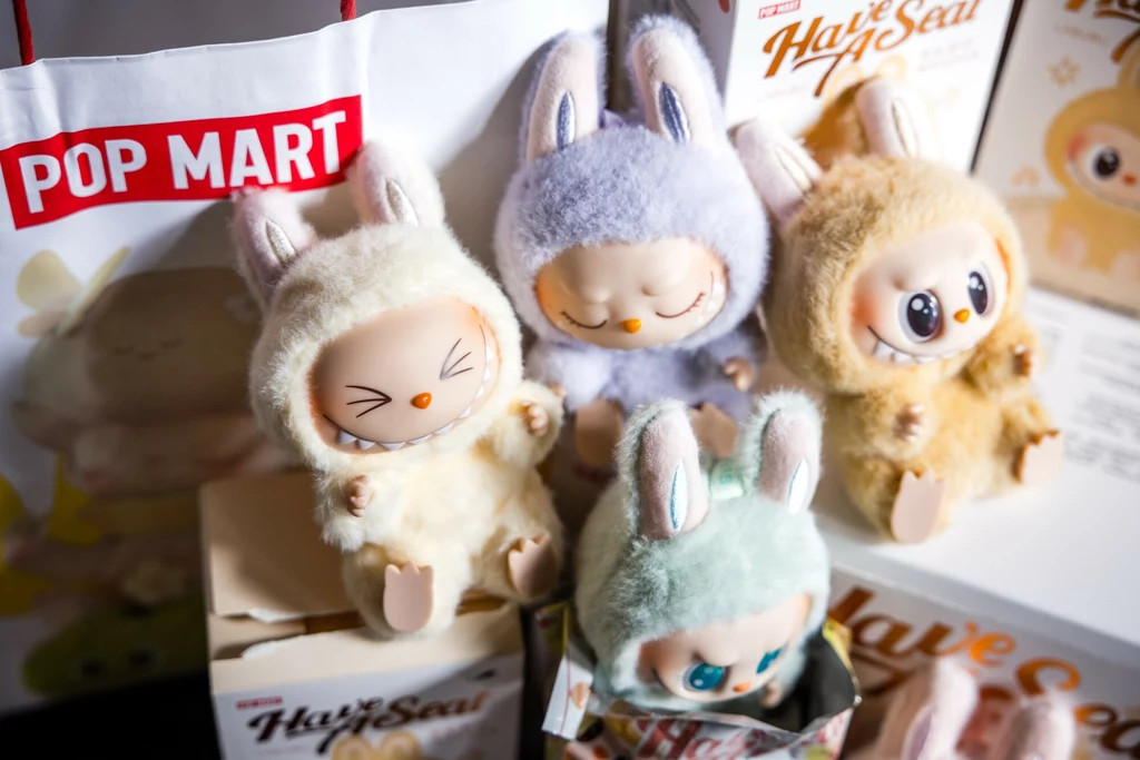 Cztery urocze figurki kolekcjonerskie z uszami królików ubrane w futerkowe kombinezony stoją obok siebie na tle opakowań i torebki z logo Pop Mart, każda z nich ma indywidualny wyraz twarzy.