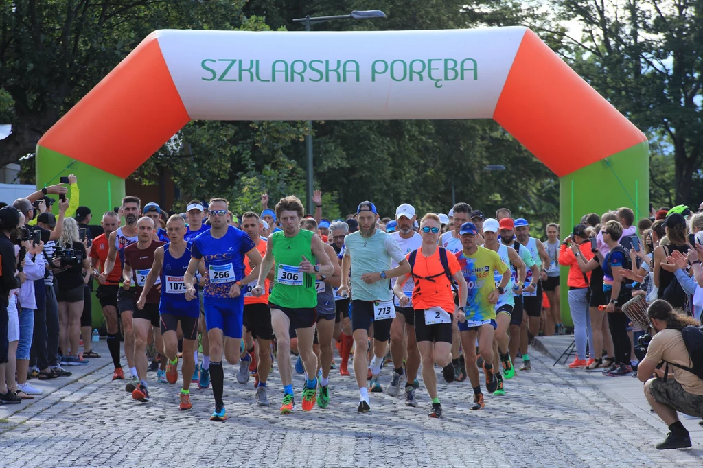 Ultramaraton w Szklarskiej Porębie Grupa biegaczy rusza spod dmuchanej bramy oznaczonej napisem Szklarska Poręba, wokół zgromadzona jest publiczność kibicująca uczestnikom biegu, całość utrzymana w atmosferze sportowego wydarzenia na świeżym powietrzu.