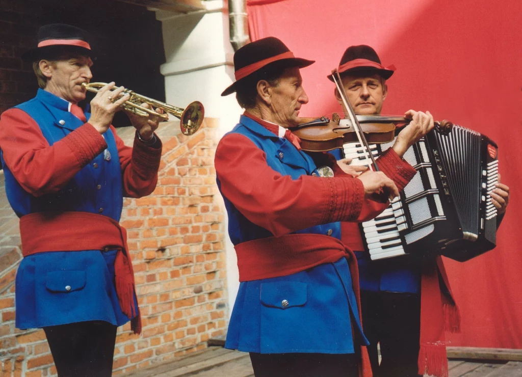 Trzech muzyków w tradycyjnych strojach ludowych gra na instrumentach: trąbce, skrzypcach i akordeonie na tle ceglanej ściany i czerwonej kotary.