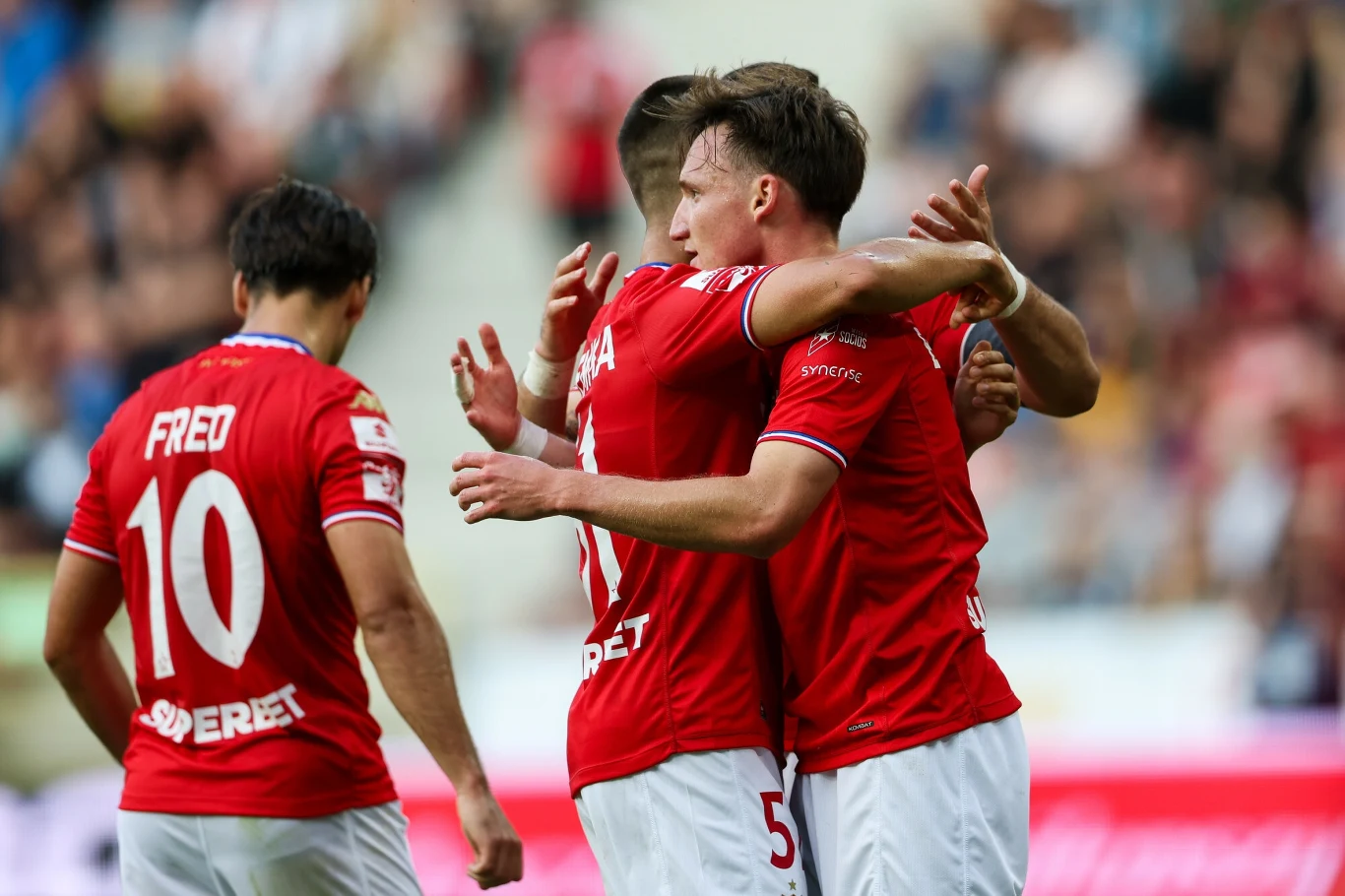 Frederico Duarte, Maciej Kuziemka, Julius Ertlthaler Piłkarze w czerwonych koszulkach przytulają się, ciesząc się wspólnie z sukcesu podczas meczu, w tle rozmyta publiczność na trybunach.