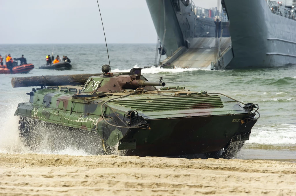Opancerzony pojazd wojskowy typu transporter opancerzony wyjeżdża na plażę z rampy dużego okrętu desantowego, w tle morze oraz ponton z żołnierzami.