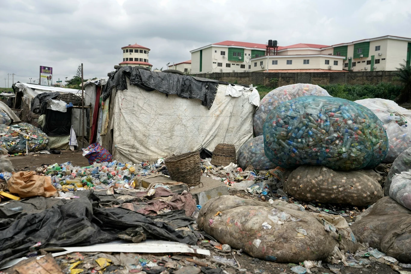 Przy prowizorycznych zabudowaniach leżą hałdy odpadów, w tym duże worki wypełnione plastikowymi butelkami, plastik i inne śmieci rozrzucone na ziemi, w tle nowoczesne budynki mieszkalne.