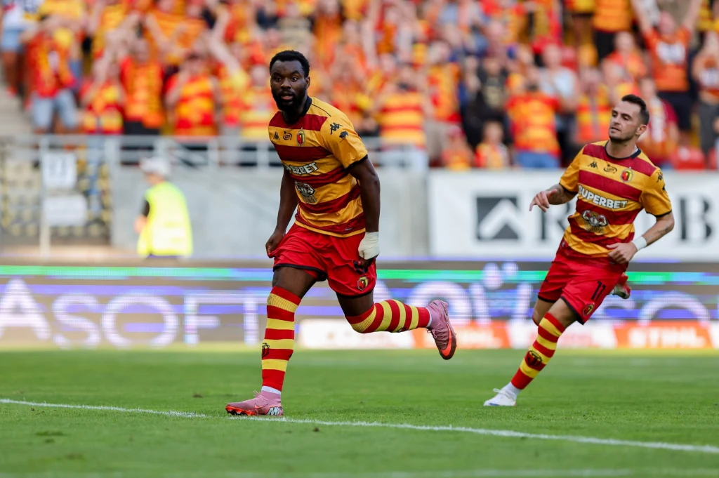 Afimico Pululu i Jesus Imaz w barwach Jagiellonii Białystok Dwóch piłkarzy w żółto-czerwonych strojach drużyny Jagiellonia Białystok biegnie po boisku podczas meczu, w tle trybuny wypełnione kibicami w barwach klubowych.