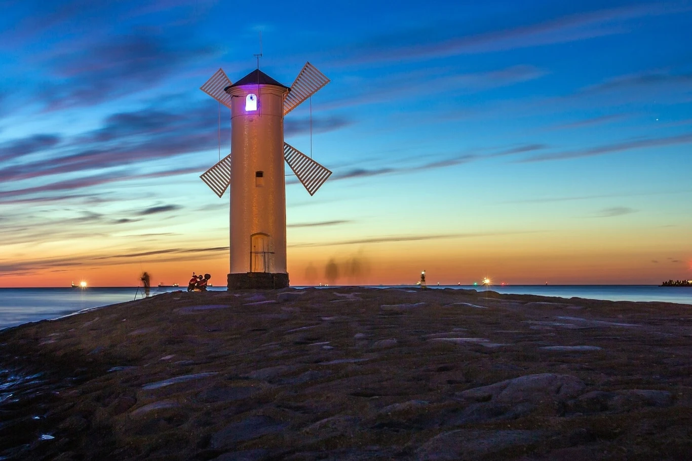 Świnoujście o zmierzchu Biała latarnia morska z charakterystycznymi skrzydłami wiatraka ustawiona na skalistym cyplu, otoczona spokojnym morzem podczas kolorowego zachodu słońca