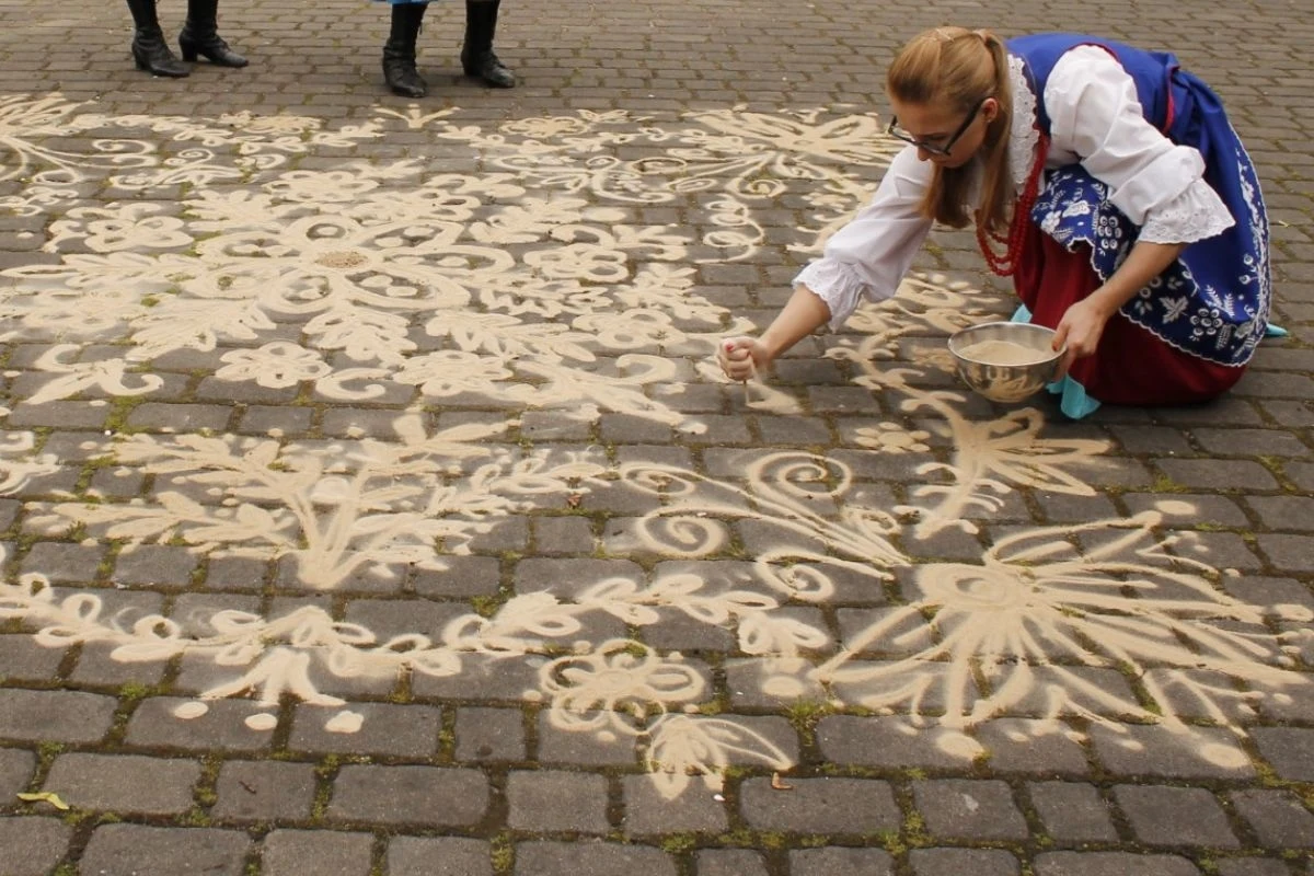 Kobieta w tradycyjnym stroju ludowym tworzy wzory na bruku za pomocą sypania piasku z miski, wokół niej rozciągające się, skomplikowane ornamenty na tle kostki brukowej, w tle widoczne tylko nogi kilku osób.