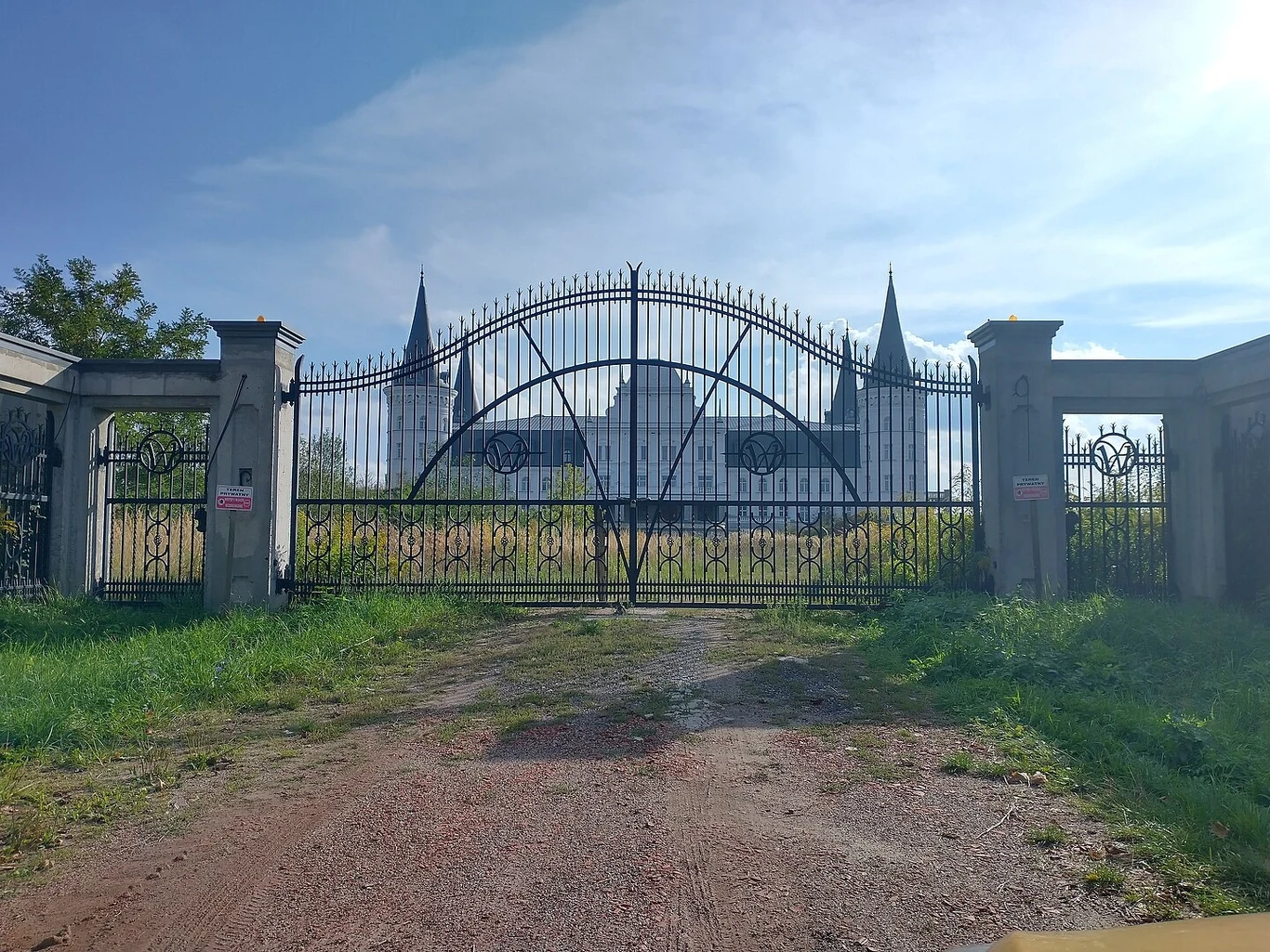 Kutrowane żelazne bramy z dekoracyjnymi elementami zamykają wjazd na ogrodzony teren, za którym widoczne są wieże budynku przypominające zamek. Po obu stronach bramy znajdują się kamienne słupy ogrodzenia oraz tabliczki ostrzegawcze, wokół teren jest z...