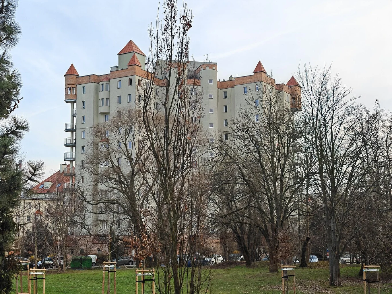 Nowoczesny budynek mieszkalny z wieloma piętrami i charakterystycznymi dachami w formie wieżyczek, otoczony bezlistnymi drzewami, na pierwszym planie widoczny zielony trawnik i fragment parku.