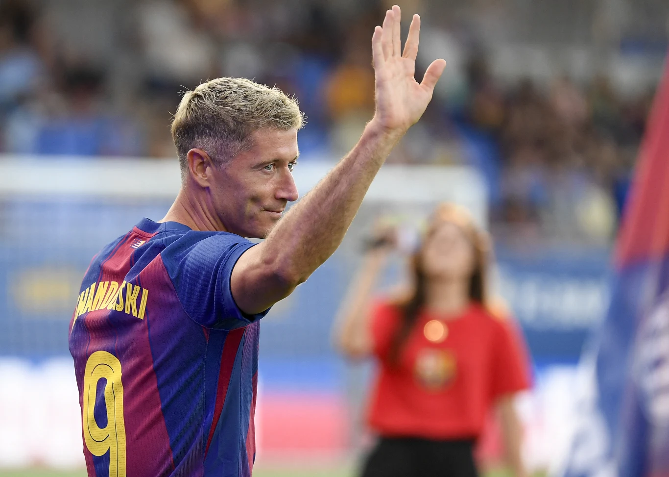 Piłkarz w stroju klubu FC Barcelona z numerem 9 na plecach macha ręką do kibiców podczas oficjalnej prezentacji na stadionie, w tle znajduje się kobieta z mikrofonem i publiczność.
