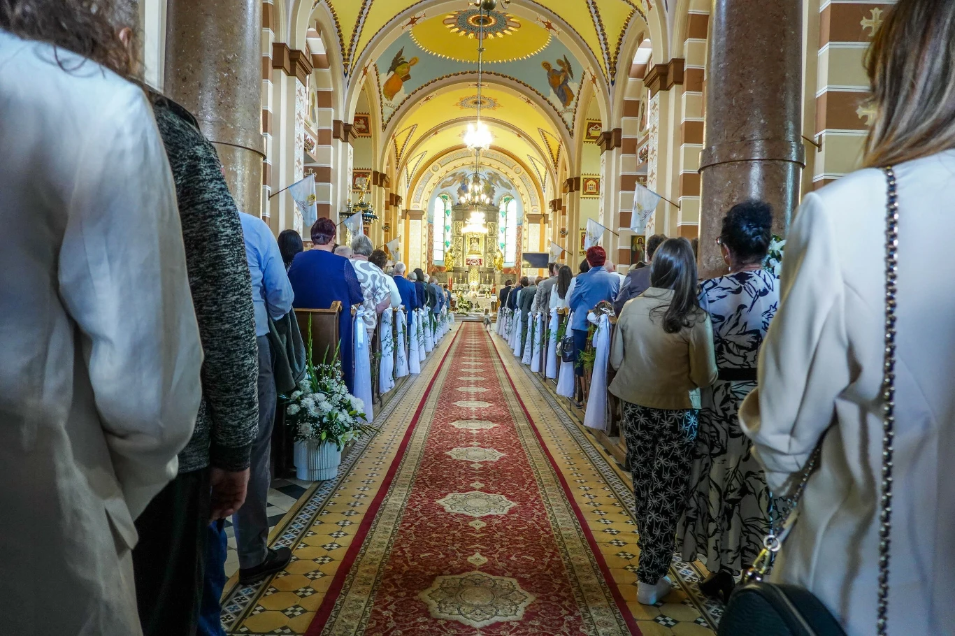Matka Boża Częstochowska. Jak zmieniała się ikona i kult, który dziś zna cała Polska? Wnętrze kościoła z licznymi wiernymi ustawionymi wzdłuż nawy, ozdobione ławki i kwiaty prowadzące do ołtarza, jasne światło wpadające przez witraże, atmosfera skupienia i uroczystości.