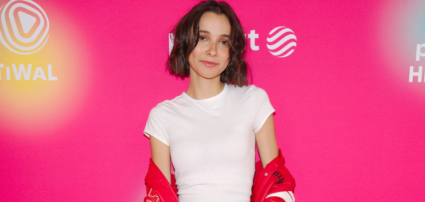 Carla Fernandes Młoda kobieta stojąca na tle jaskrawego, różowego bannera z białymi napisami. Ubrana w białą koszulkę i rozpiętą czerwoną kurtkę, uśmiecha się lekko do obiektywu.