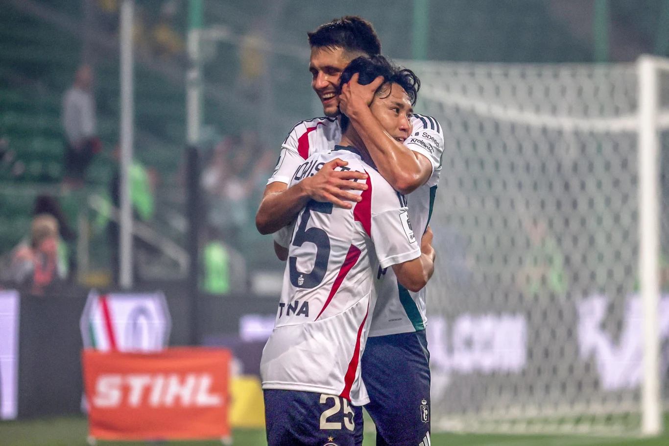 W ostatniej kolejce Legia pokonała GKS Katowice, teraz musi odrobić straty z meczu z AEK Larnaka Dwóch piłkarzy w biało-czerwonych strojach obejmuje się na murawie stadionu, okazując radość i emocje po zdobyciu bramki lub zwycięstwie, w tle bramka oraz rozmyte sylwetki kibiców.