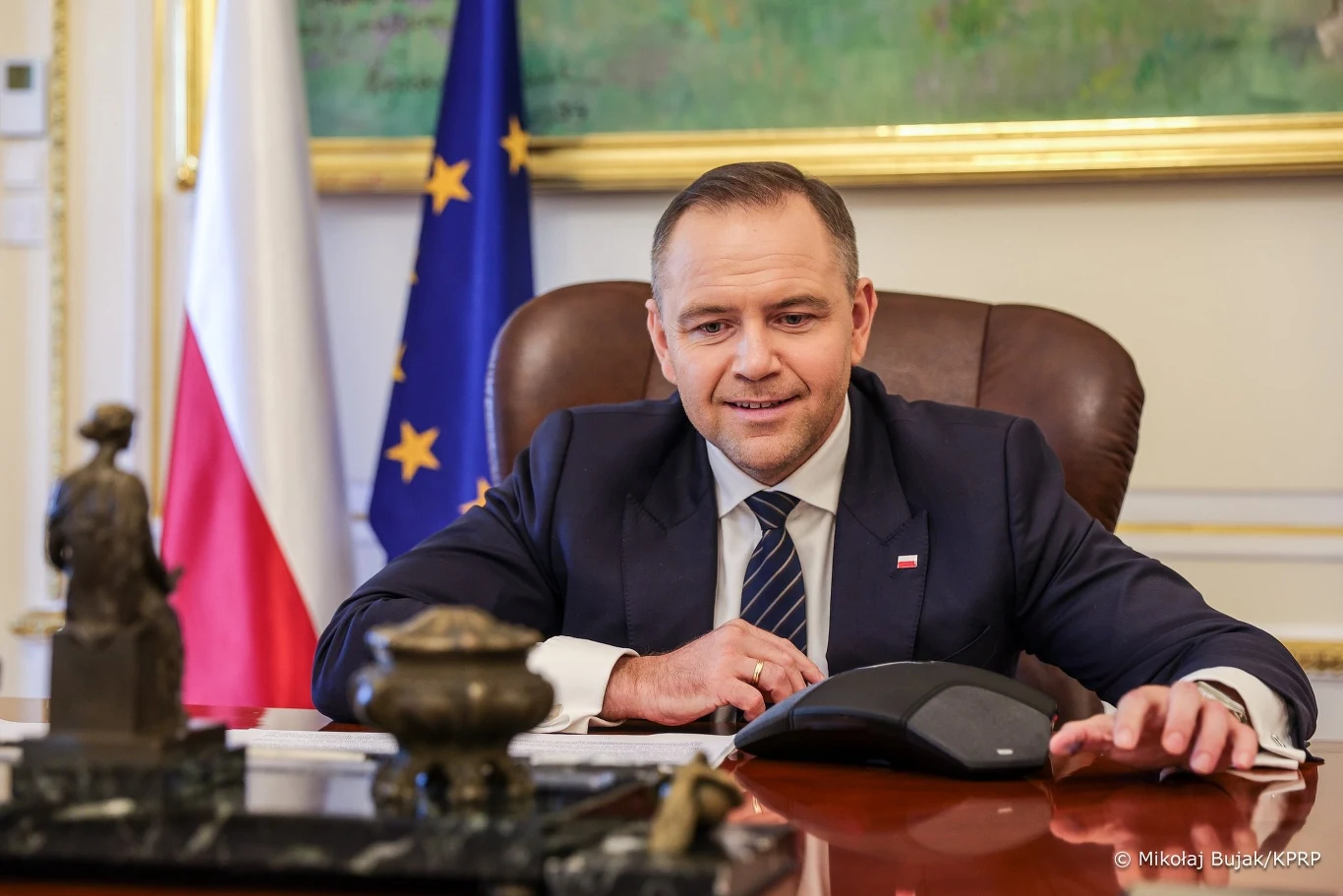 Karol Nawrocki brał udział w telekonferencji z Donaldem Trumpem Mężczyzna w garniturze siedzi przy biurku w eleganckim gabinecie, za nim znajdują się flagi Polski oraz Unii Europejskiej, na biurku widoczne są dokumenty oraz ozdobna figurka.
