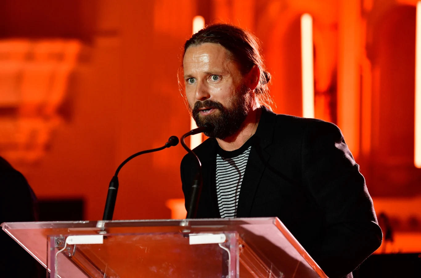 Max Martin Mężczyzna z brodą, ubrany w czarną marynarkę i pasiastą koszulkę, stoi za przezroczystą mównicą podczas wydarzenia na tle pomarańczowego oświetlenia.