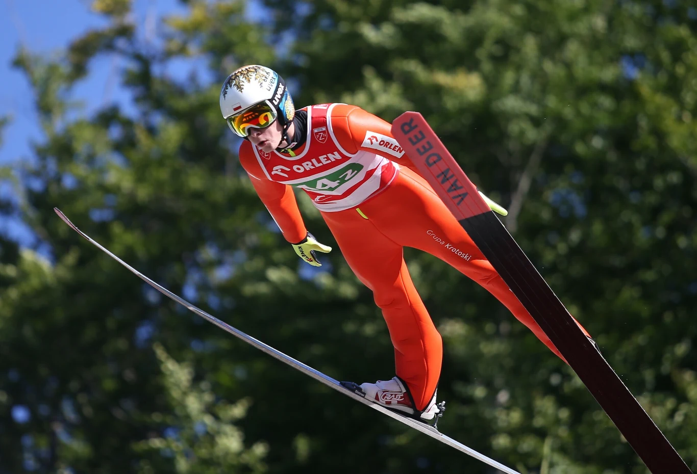 Skoczek narciarski w czerwonym kombinezonie leci w powietrzu podczas zawodów, w tle rozmyte zielone drzewa i niebieskie niebo, widoczny napis Van Deer na jednej z nart oraz logo Orlen na stroju sportowca.