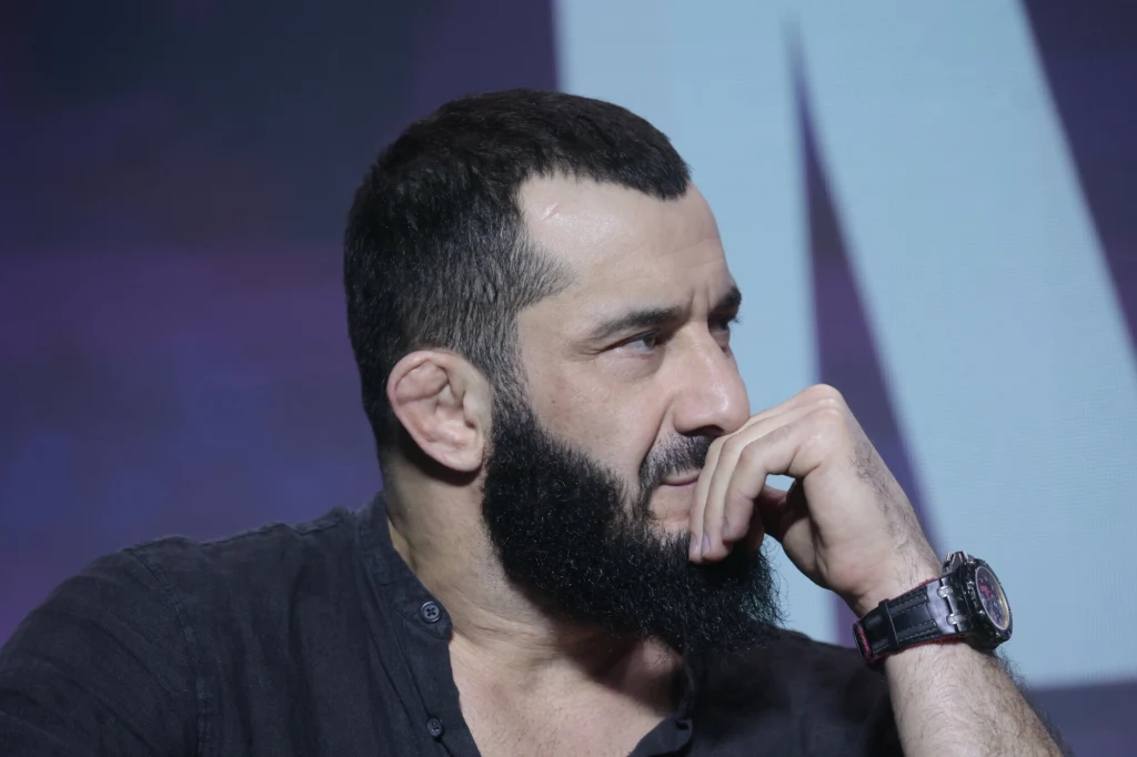 Mężczyzna z krótkimi, ciemnymi włosami oraz gęstą, czarną brodą siedzi z zamyśloną miną, opierając dłoń na ustach; ubrany w ciemną koszulę, na nadgarstku nosi zegarek; w tle rozmazane, fioletowo-niebieskie tło.