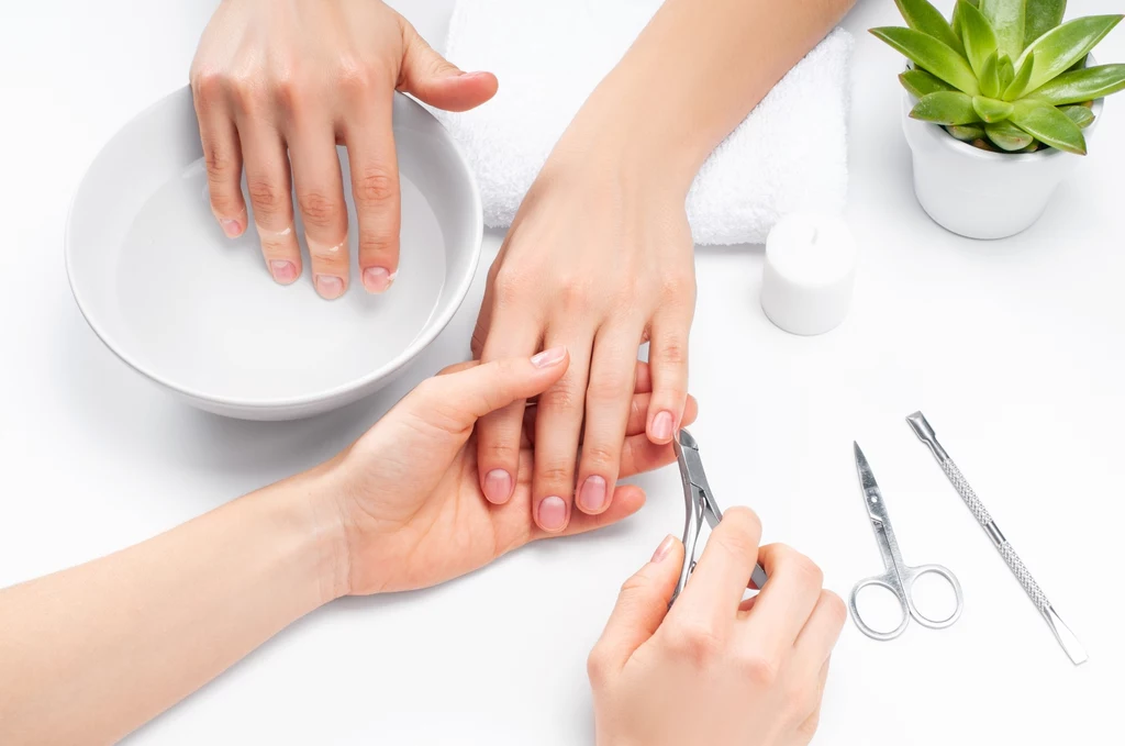 Ręce osoby podczas zabiegu manicure, jedna dłoń moczona w miseczce z wodą, druga przygotowywana przez stylistkę przy użyciu cążek do skórek, obok leżą narzędzia kosmetyczne oraz doniczka z zieloną rośliną.