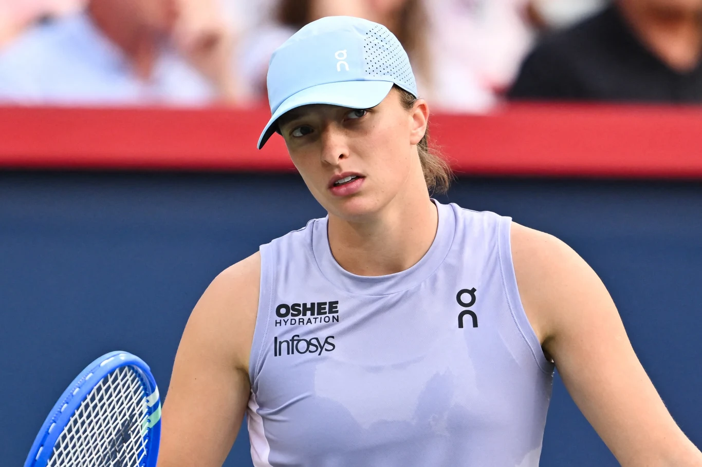 Iga Świątek Tenisistka w jasnej koszulce oraz czapce z daszkiem trzyma rakietę tenisową na tle rozmytej publiczności, widoczne wyraźne logotypy sponsorów na jej stroju.