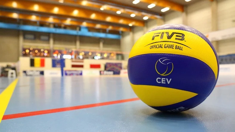 Kolorowa piłka siatkarska FIVB z widocznym logo CEV umieszczona na błyszczącej hali sportowej, w tle rozmyte trybuny oraz flagi państwowe.