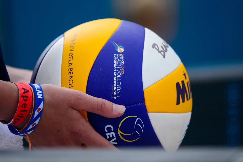 Ręka zawodnika trzymająca piłkę do siatkówki plażowej z widocznym logotypem zawodów oraz kolorową opaską na nadgarstku.