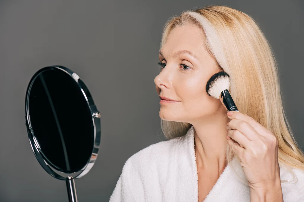 Czym jest soft-sculpted make-up? Co to za makijaż? Kobieta o blond włosach w białym szlafroku nakłada makijaż za pomocą pędzla, patrząc w okrągłe lustro ustawione przed sobą na tle jednolitej, szarej ściany.
