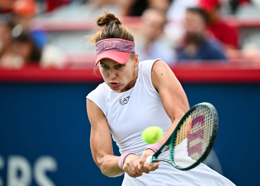 Weronika Kudiermietowa Zawodniczka tenisa w białym stroju sportowym skupiona na odbiciu piłki rakietą podczas meczu, dynamiczny ruch i pełna koncentracja na twarzy, w tle rozmyta publiczność.