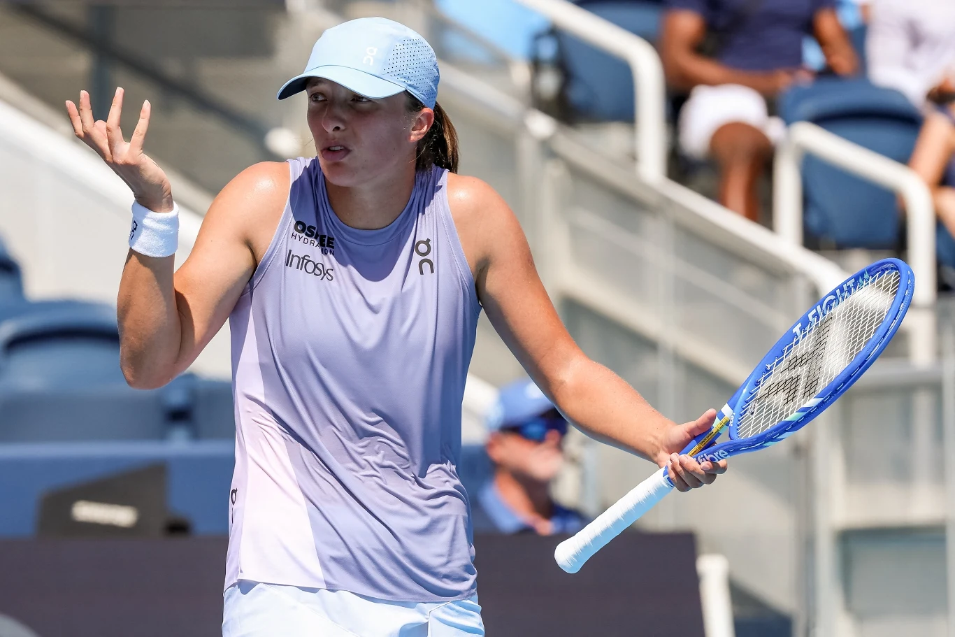 Iga Świątek Zawodniczka tenisa w sportowym stroju i czapce z daszkiem stoi na korcie, trzyma rakietę tenisową w jednej ręce, a drugą wykonuje gest zdziwienia lub niezrozumienia; w tle widoczne trybuny.