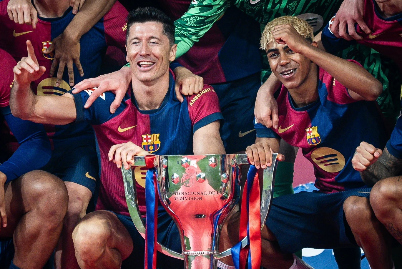 Lamine Yamal (z prawej) i Robert Lewandowski Piłkarze drużyny FC Barcelona w strojach klubowych świętują zdobycie trofeum, skupieni wokół dużego pucharu z napisem La Liga, wyrażają radość i dumę z osiągniętego sukcesu.