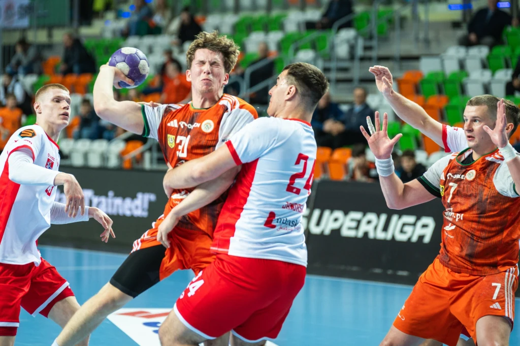 Mecz Azotów Puławy z MKS Zagłębiem Lubin w sezonie 2024/2025 Emocjonujący moment podczas meczu piłki ręcznej, zawodnik w pomarańczowej koszulce próbuje oddać rzut, będąc blokowanym przez przeciwnika w biało-czerwonym stroju, a pozostali zawodnicy aktywnie uczestniczą w akcji na tle hali sportowej.
