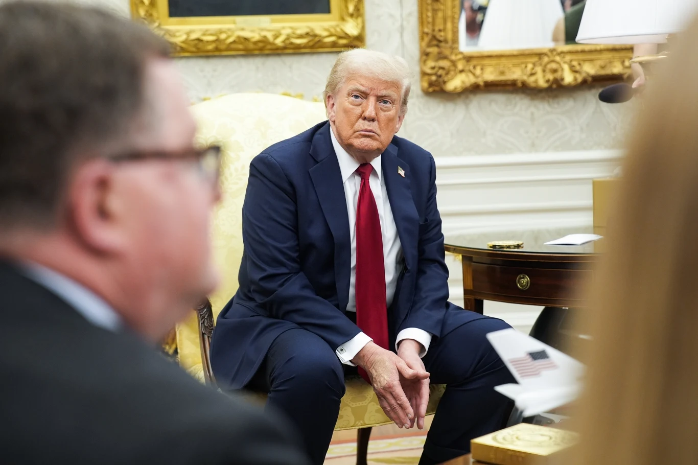 Prezydent USA Donald Trump Mężczyzna w garniturze z czerwoną krawatą siedzi na jasnym fotelu w eleganckim, oficjalnym gabinecie, wokół niego znajdują się inne osoby, widoczne są ozdobne ramy obrazów i biurko.