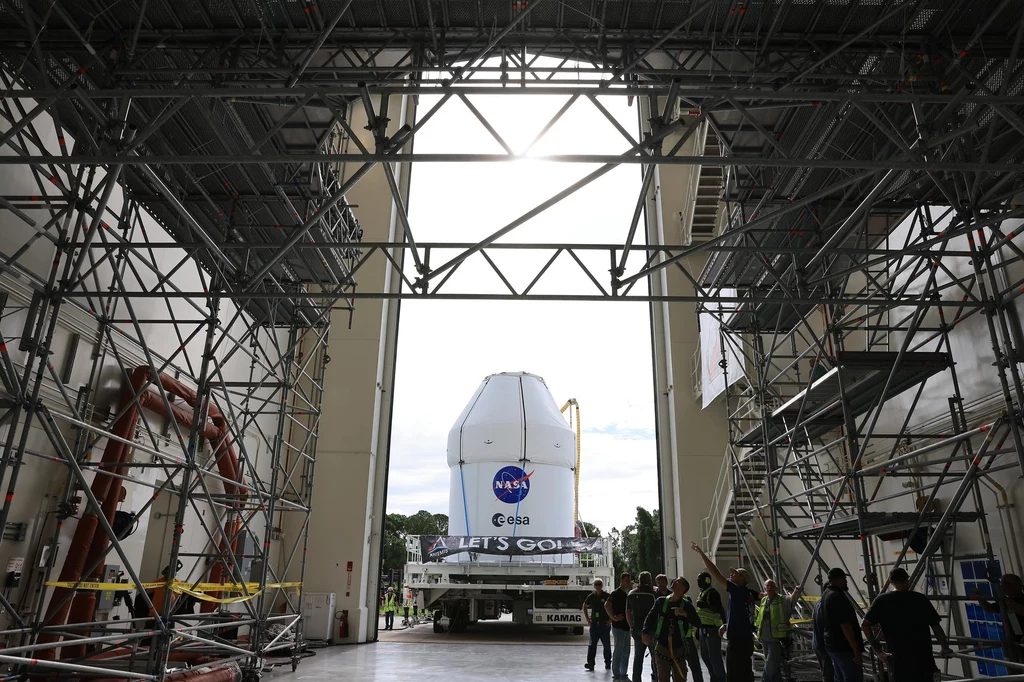 Kapsuła załogowa Orion dla misji Artemis 2. NASA prowadzi testy statku w centrum na Florydzie. Duża kapsuła kosmiczna NASA transportowana na specjalnej platformie przez otwarty hangar, wokół konstrukcje stalowe i grupa pracowników obserwujących przemieszczanie ładunku.