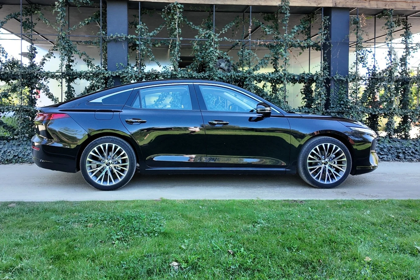 Czarny nowoczesny samochód typu sedan stoi na chodniku na tle zielonej ściany z pnączami i oknami. Auto ma elegancką sylwetkę, duże aluminiowe felgi i lśniącą karoserię.