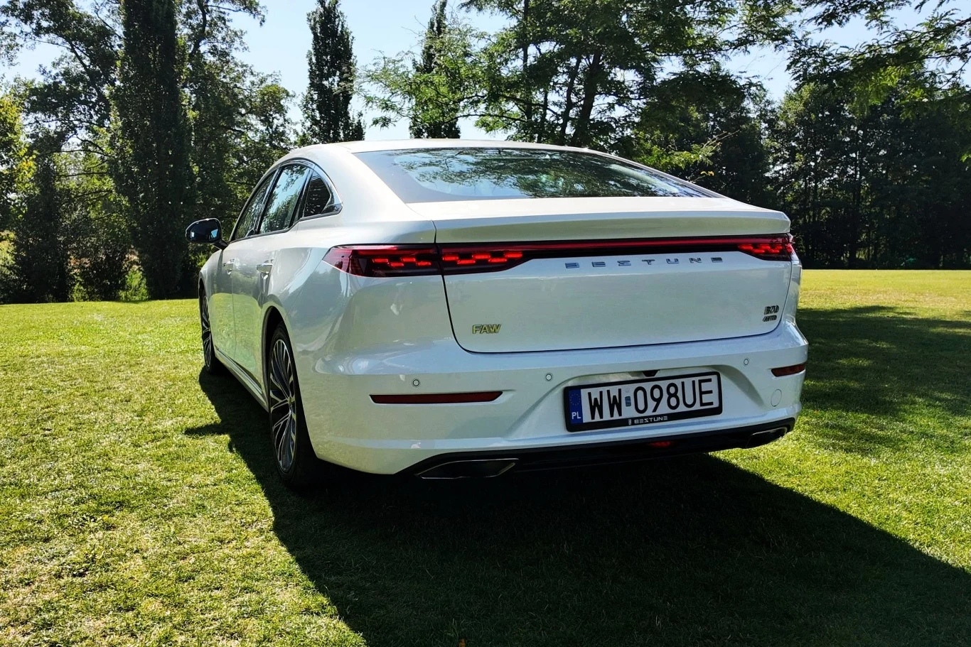 Biały nowoczesny samochód sedan marki Bestune stoi na zielonej trawie w słoneczny dzień, na tle drzew; widoczny jest tylni zderzak, lampy LED oraz tablica rejestracyjna.