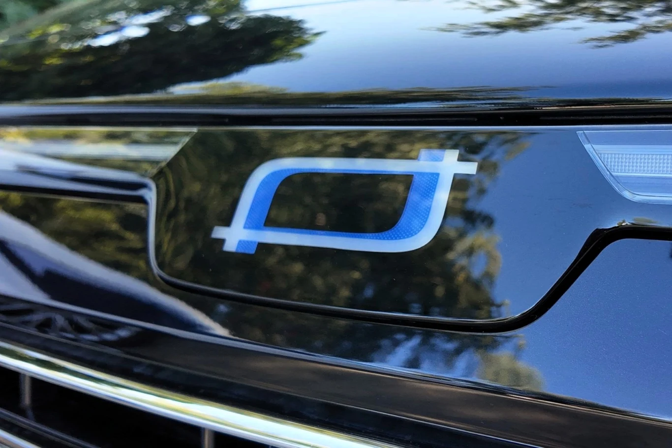 Błyszczący fragment przodu samochodu z wyraźnym niebieskim logotypem motoryzacyjnym na ciemnym tle, odbijającym otaczającą zieleń.